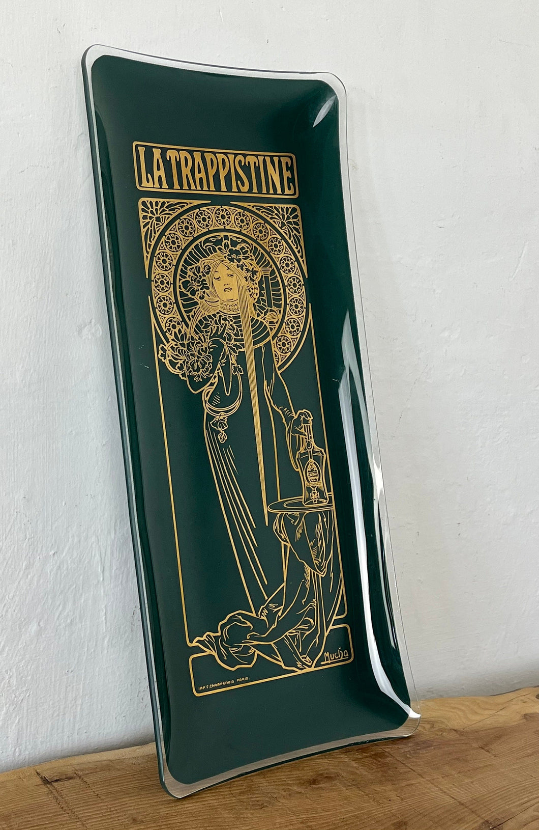 The picture shows a vintage glass tray with deep green background and Alphonse Mucha's painting La Trapisttine in gold. Depicts a young woman dressed in a religious habit with a calm and serene expression. One of her hands is on a bottle.