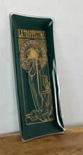 Load image into Gallery viewer, The picture shows a vintage glass tray with deep green background and Alphonse Mucha&#39;s painting La Trapisttine in gold. Depicts a young woman dressed in a religious habit with a calm and serene expression. One of her hands is on a bottle.
